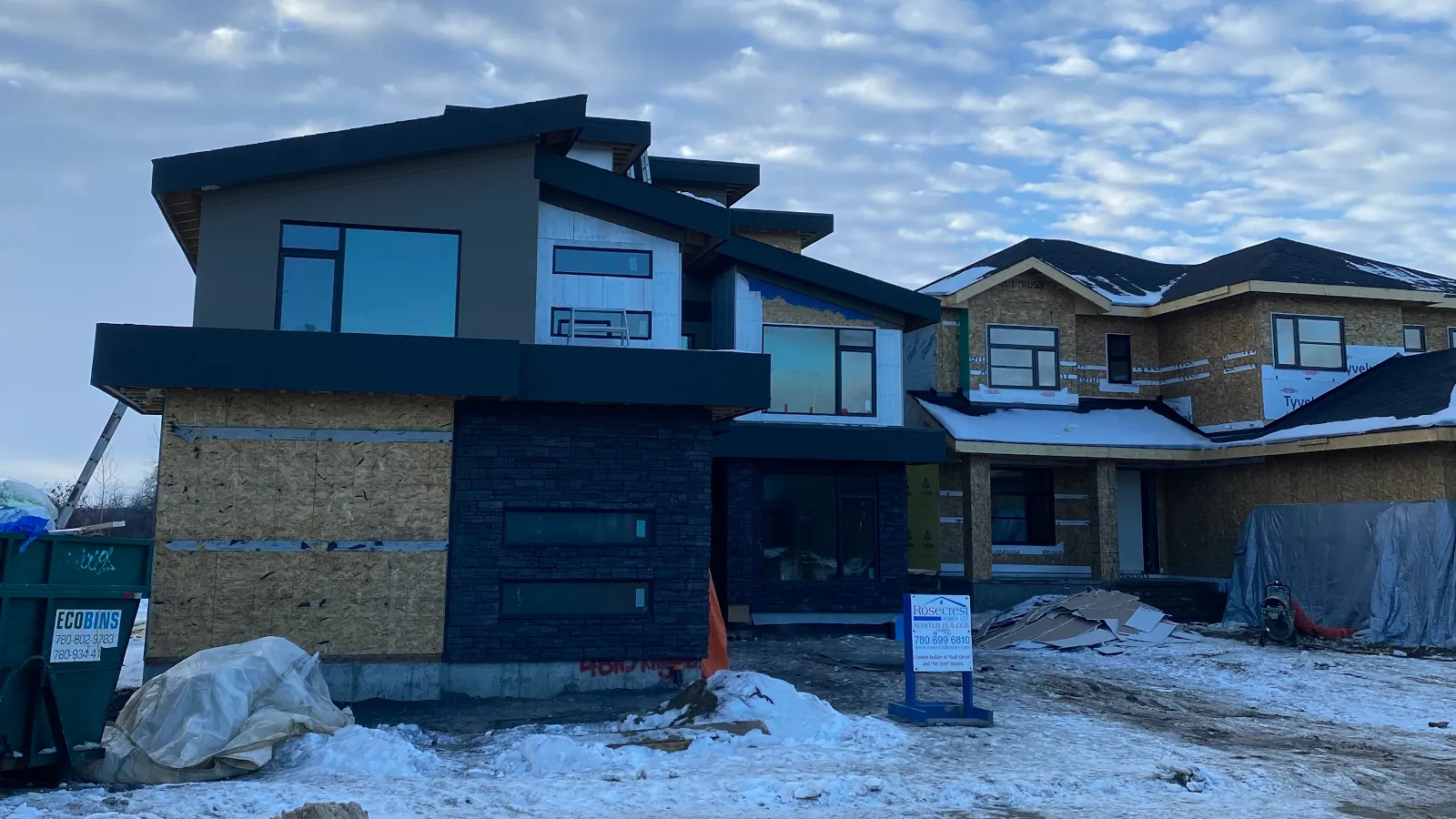 A large three-story house under construction in winter, showing finished beige stucco and culture stone alongside sections with exposed house wrap.