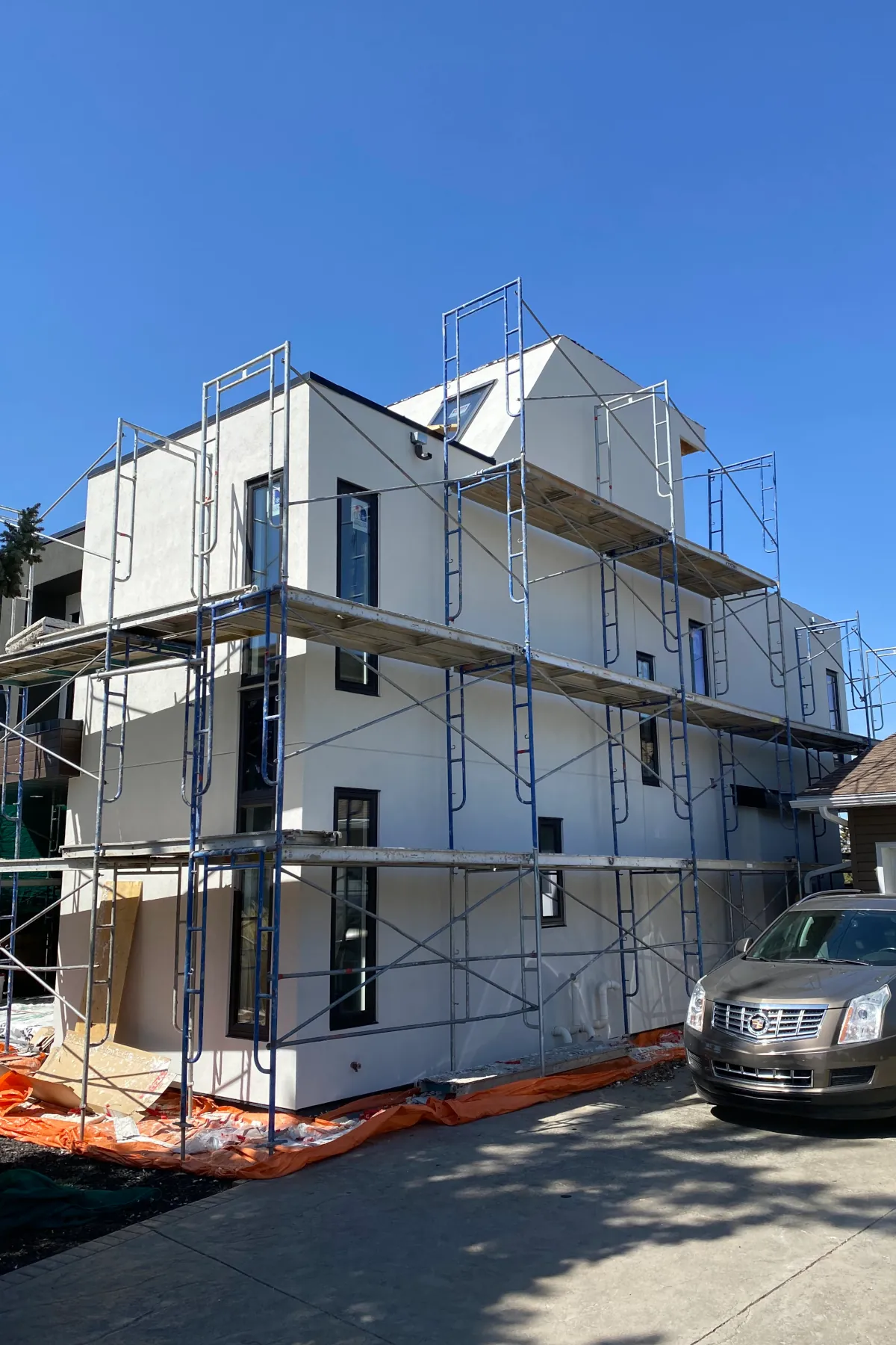 A two-story modern house in Edmonton with blue scaffolding erected for the application of the final stucco finish coat.